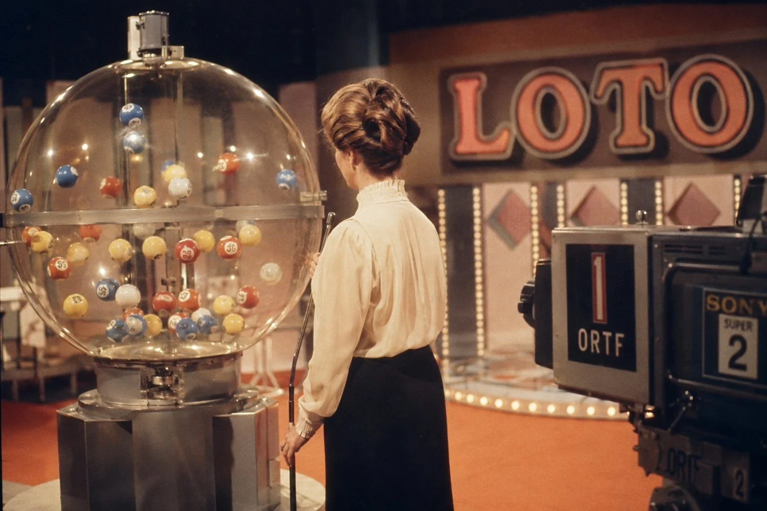Plateau de télévision française dans les années 1970, sphère transparente de tirage avec boules colorées numérotées en mouvement, présentatrice de dos en blouse à col haut, caméras de studio noires en bord de plateau, grand logo LOTO en typographie chunky